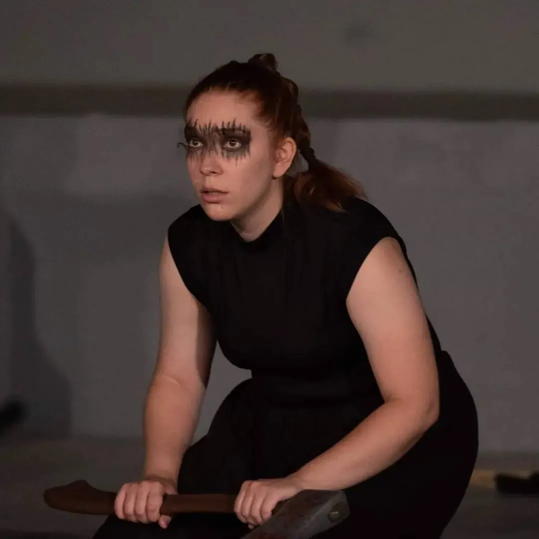 Maria on stage in a black dress with dramatic make-up, holding a small axe