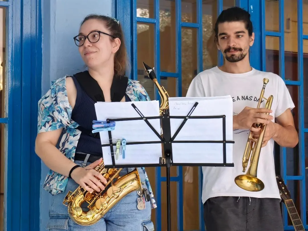 Maria on saxophone with a trumpeter playing in front of a blue gate