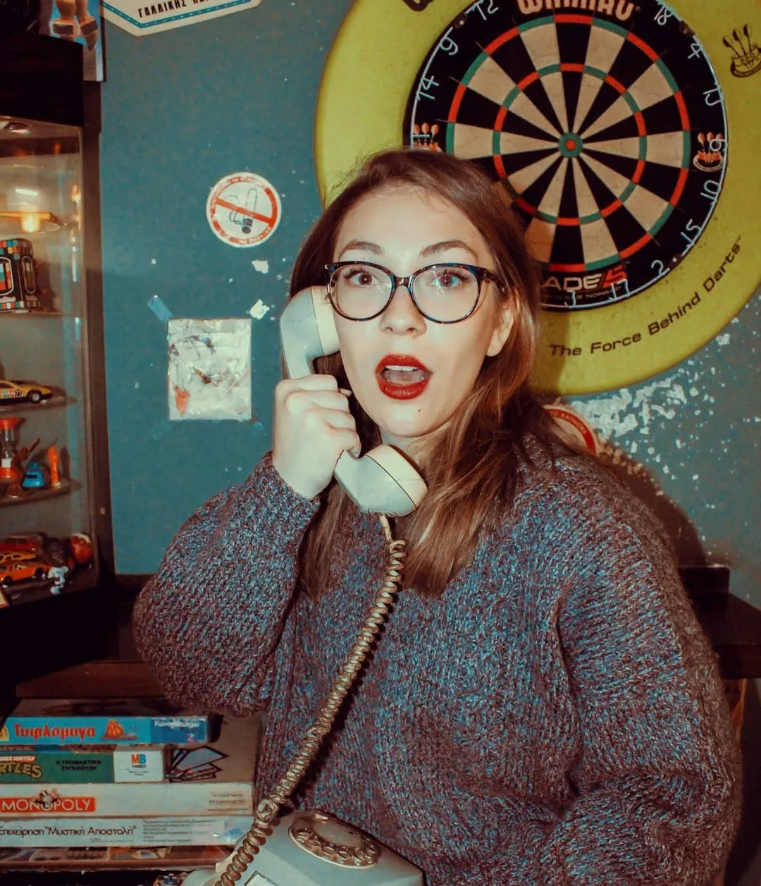 Maria in a knitted jumper on an old telephone in front of a dartboard, a comic scene