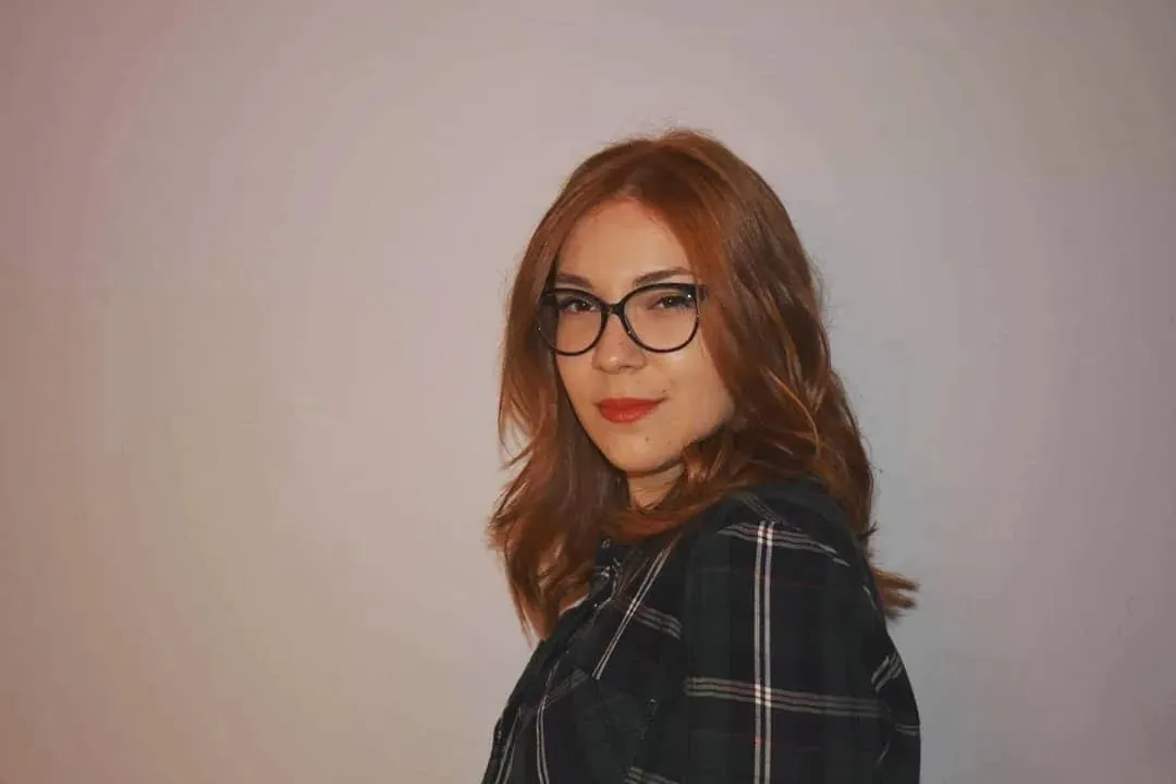 Maria with red hair and glasses in a checked shirt, looking over her shoulder