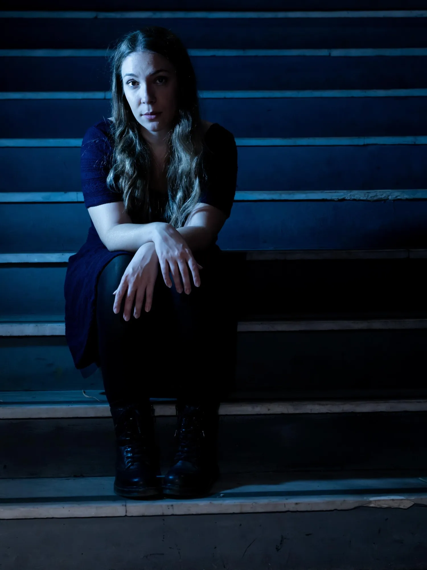 Maria sitting on dark stone steps in blue light, looking into the camera